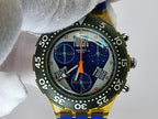 A Swatch wristwatch with a yellow-tone case, blue and gray dial, and dark green bezel, held by a white glove.