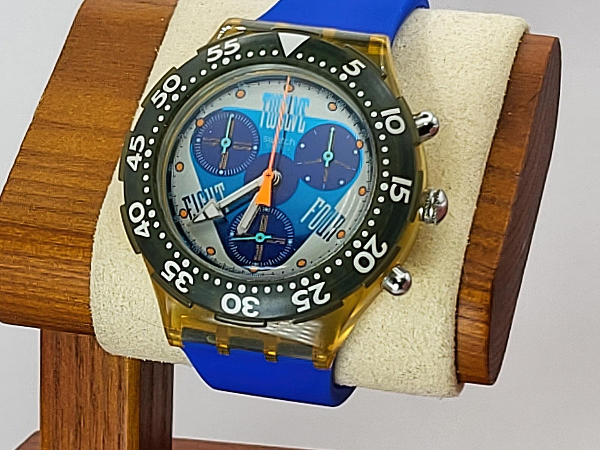 A Swatch wristwatch wristwatch with a translucent gold case, blue strap, and a blue dial featuring three subdials and a black bezel, displayed on a cream cushion inside a wooden stand.