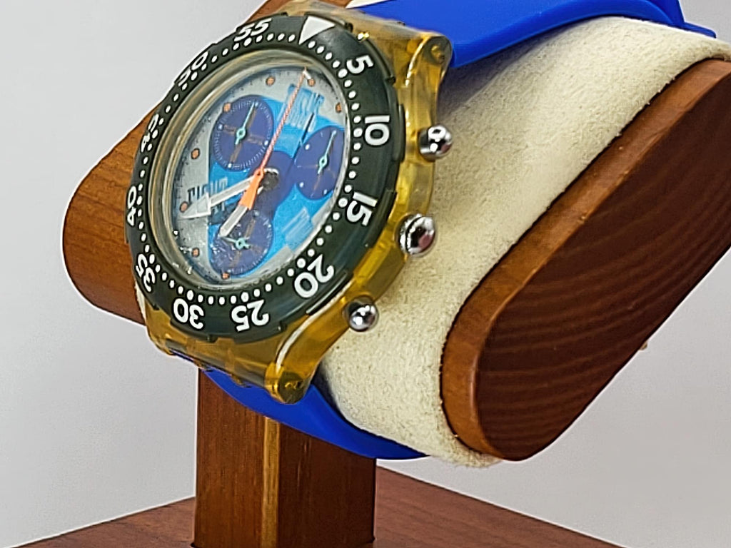 A Swatch wristwatch with a translucent amber-yellow case, blue strap, and blue dial with a black bezel, displayed on a wooden stand with a cream cushion.