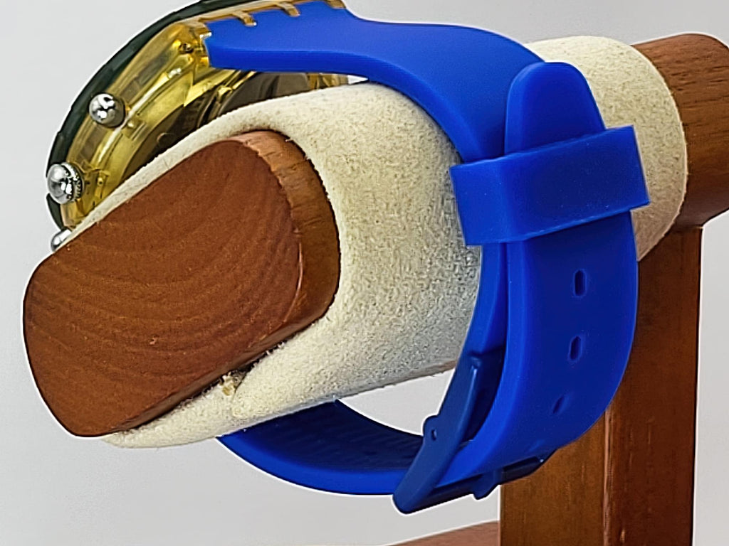 A close-up of a Swatch wristwatch with a blue silicone strap and a gold-toned transparent case, mounted on a wooden display stand with a beige foam sleeve.