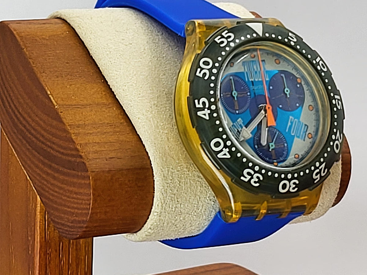 A Swatch wristwatch with a translucent amber-yellow case, blue strap, black bezel, and blue dial, displayed on a cream cushion mounted on a wooden stand.