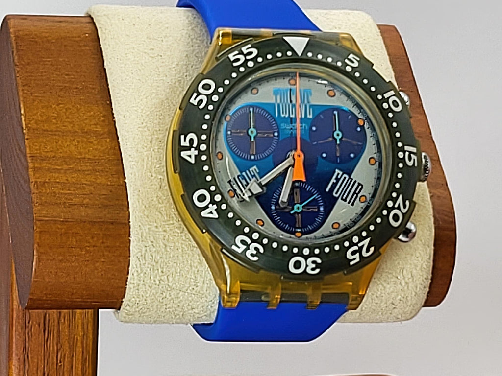 A Swatch wristwatch with a translucent amber-yellow case, blue strap, and blue dial, displayed on a cream cushion mounted on a wooden stand.