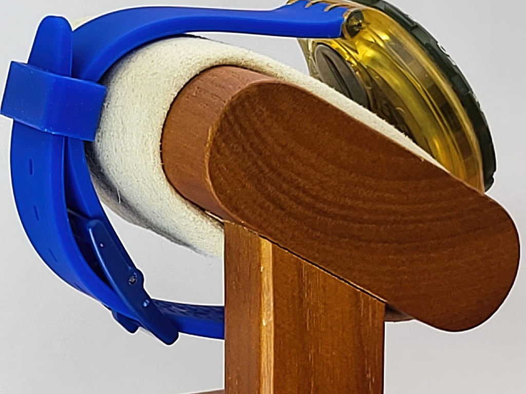 A close-up of a blue and white Swatch wristwatch mounted on a wooden display stand, with a translucent yellow circular component visible behind it against a light gray background.