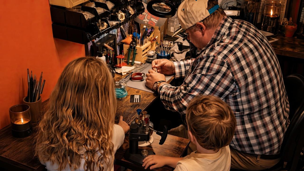 A father in a plaid shirt and cap works at a cluttered watch craft table with his two children watching closely in a warmly lit workshop.